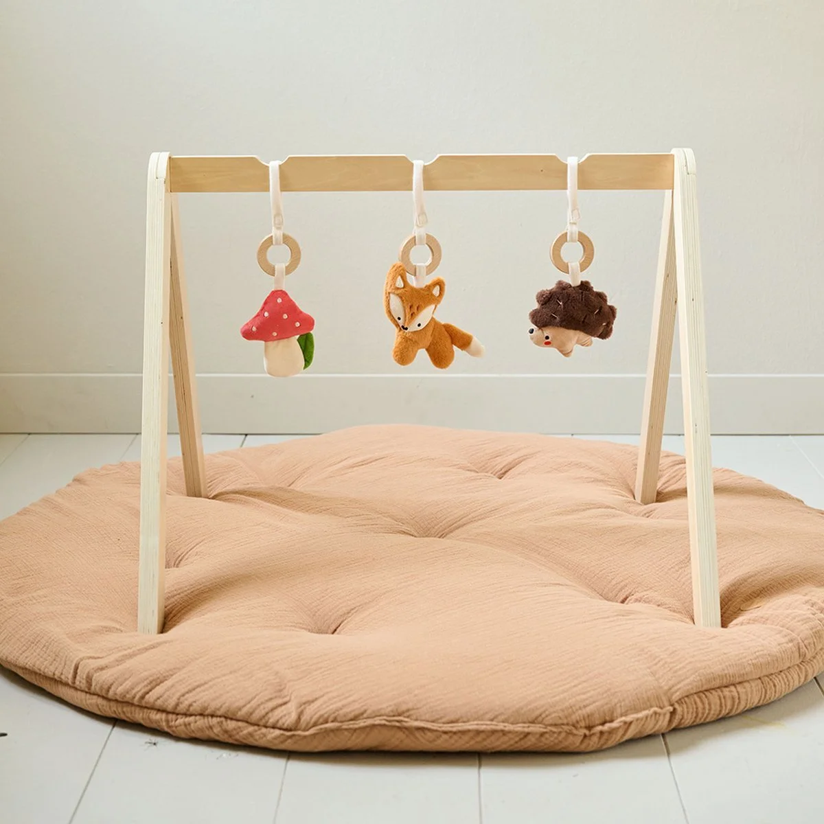 Wooden baby gym with hanging plush toys over a pink padded mat on a white floor.