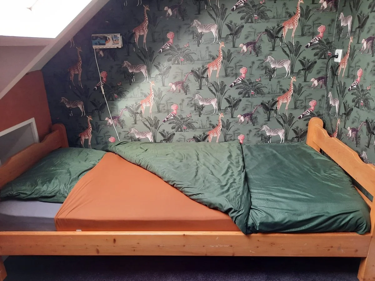 A bedroom with a bed against a jungle-themed wallpaper featuring giraffes, zebras, and other animals with green leaves. The bed has green and orange bedding.
