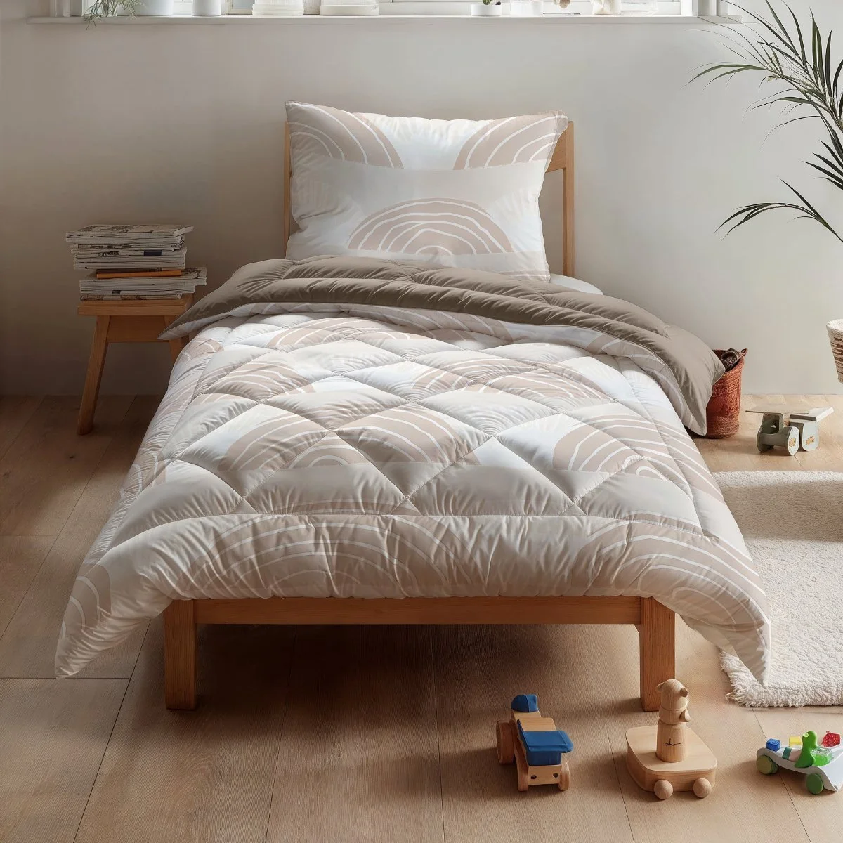 A neatly made single bed with a wooden frame, white and beige patterned bedding, and a pillow, located in a bright room with natural light. Next to the bed is a small wooden nightstand with a stack of magazines. The floor is wooden, and there are children's toys nearby, with plants and a window in the background.