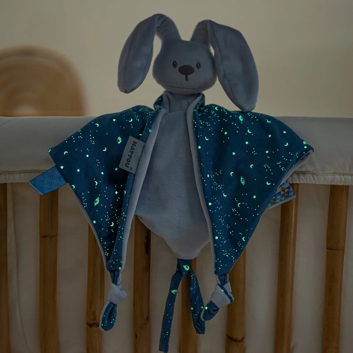 Gray plush bunny toy with a moon and stars pattern on its cape, hanging from a crib railing in a dimly lit room.