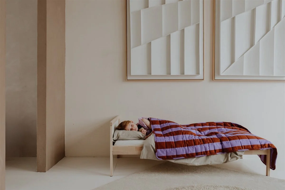 A girl lying in bed with a quilt, resting her head on a pillow, in a minimalistic, modern bedroom with framed geometric artwork on the wall.
