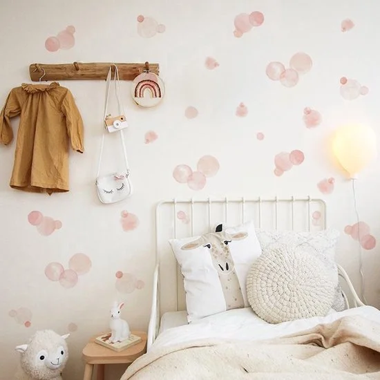 Bedroom with white bed, decorative pillows including a deer face and knitted round pillow, wall with pink and beige circular patterns, a wooden coat rack with a coat, rainbow wall hanging, and crossbody bag, nightstand with a rabbit figurine, and a floor lamp shaped like a balloon.