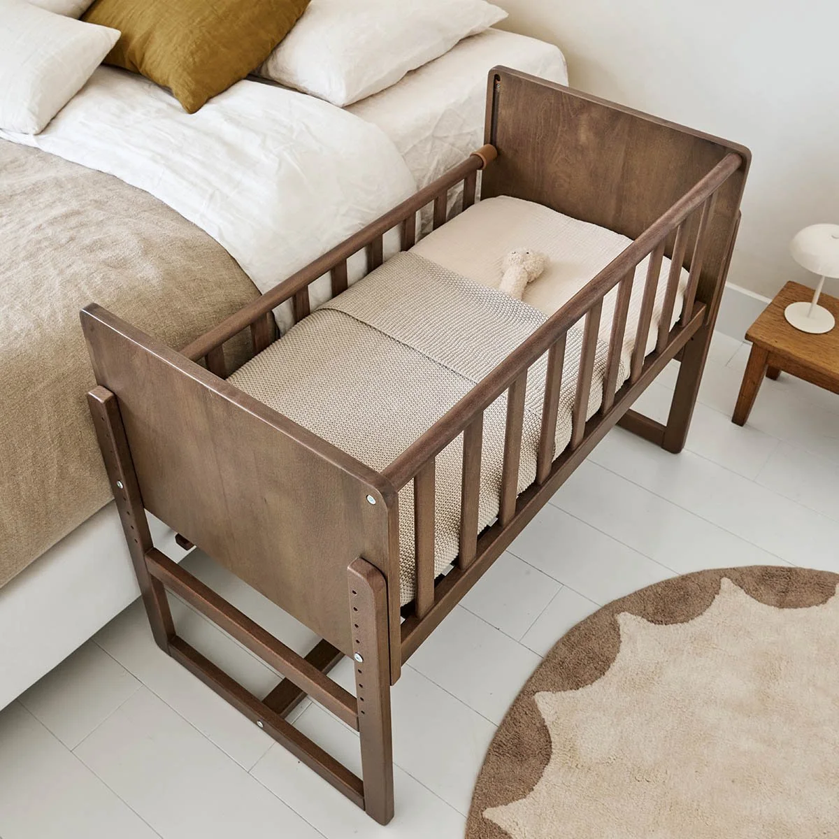 A wooden crib with a beige mattress and a small stuffed toy inside, next to a bed with beige and white bedding, and a small wooden side table with a white lamp on it, on a white floor with a beige and brown round rug.