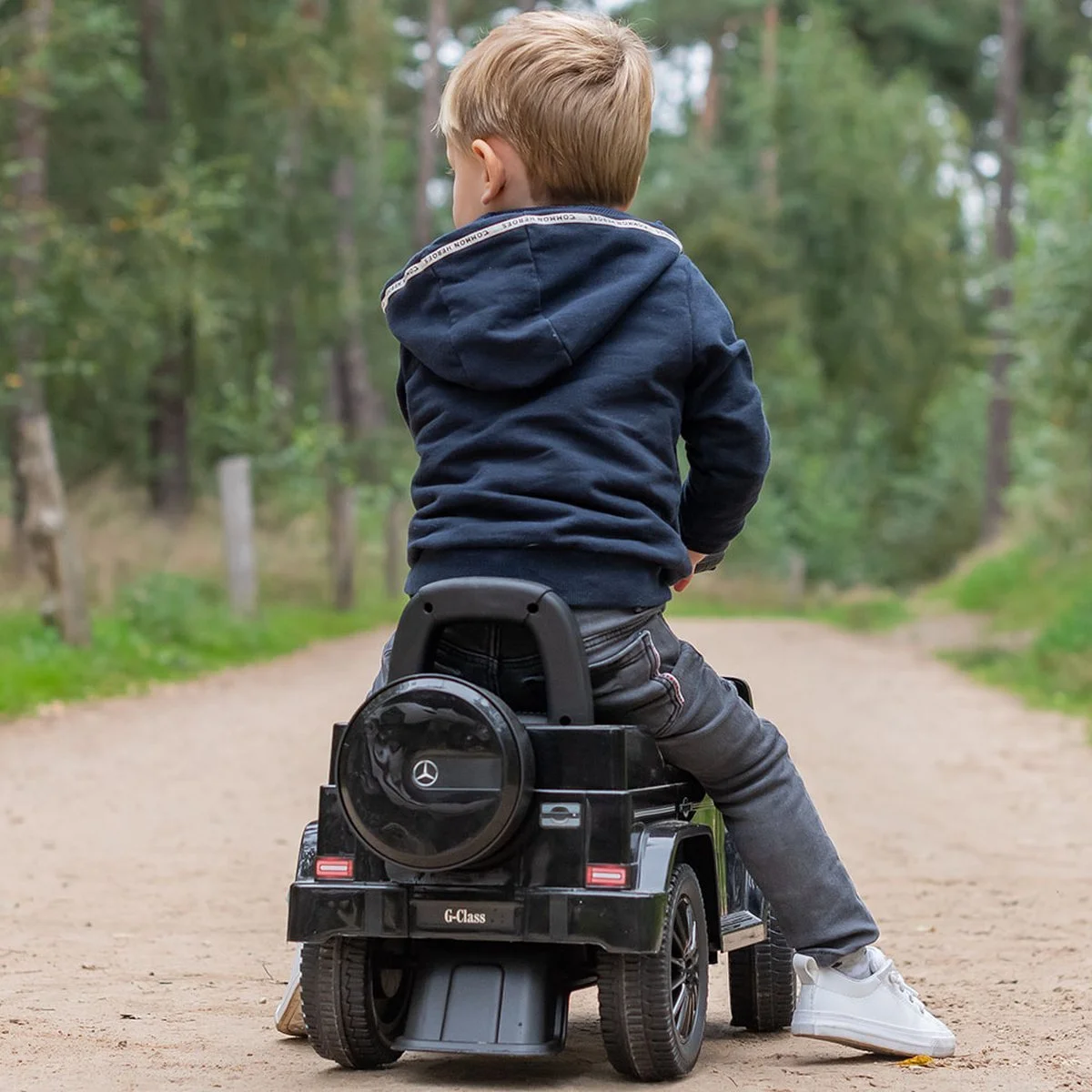Child riding a toy car on a dirt path in a wooded area, viewed from behind. The boy wears a navy hoodie, gray pants, and white sneakers.
