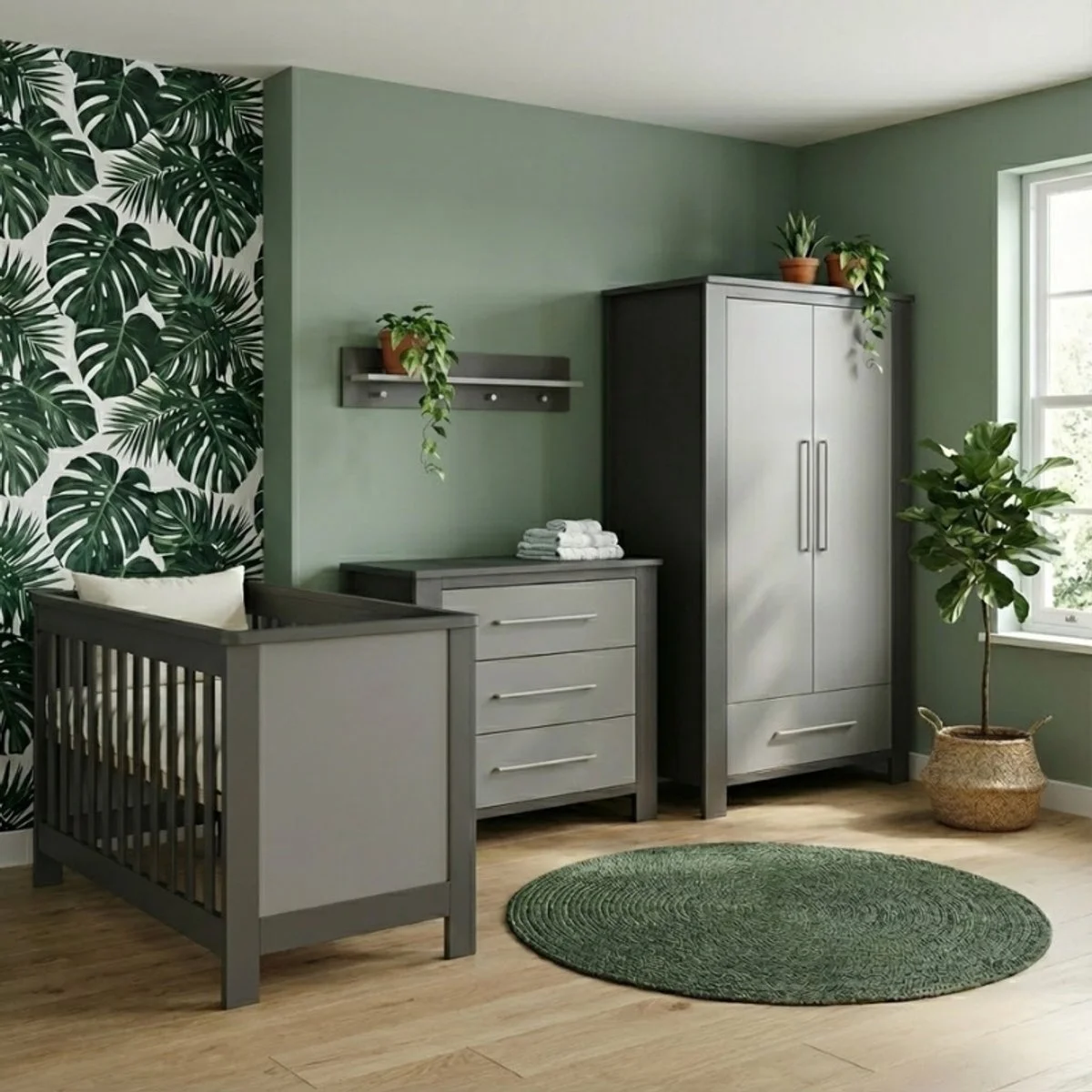 Nursery room with green leafy wallpaper on one wall, gray furniture including a crib, dresser, and wardrobe, a green circular rug, potted plants, and a wicker basket in the corner near a window.