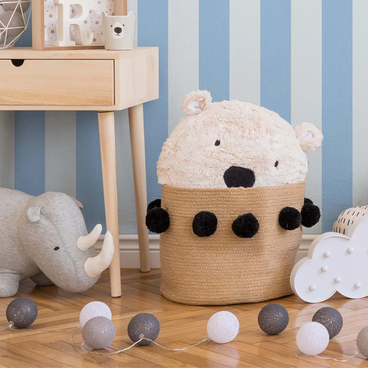 Decorative plush toys in a nursery with a large bear in a woven basket, a small elephant, and a cloud-shaped light, against a blue striped wall. String lights with gray and white balls are on the wooden floor.