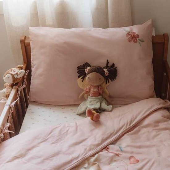 A child's bed with light pink sheets and pillowcase, decorated with embroidered flowers and hearts, with a plush doll sitting against the pillow, and a stuffed animal hanging over the side of the bed.