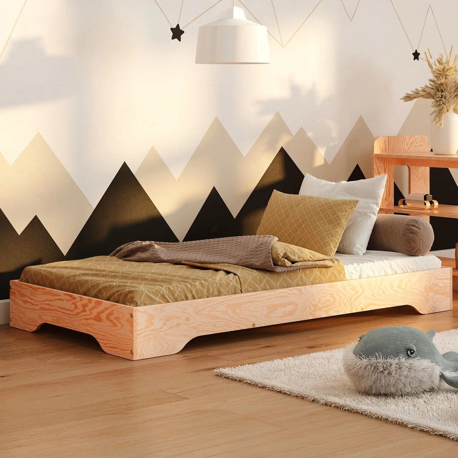 A bedroom with a wooden bed frame, mustard yellow patterned bedding, a beige pillow, and a neutral bolster pillow. There is a geometric mountain mural on the wall, a white pendant light, a small wooden shelf with decorative items, a vase with dried wheat, and a plush whale-shaped rug on the wooden floor.