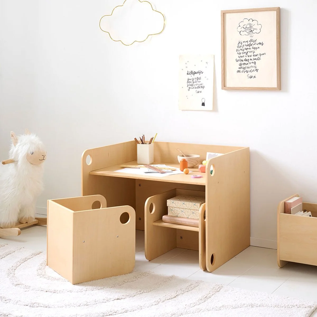 A children's play area with a light wooden table and matching chairs, decorated with colorful toys and art supplies. There is a plush llama on a rocking base on the left side, surrounded by artwork and framed drawings on the white wall above. The room has a soft, cream-colored rug and a small wooden storage box filled with books on the right.