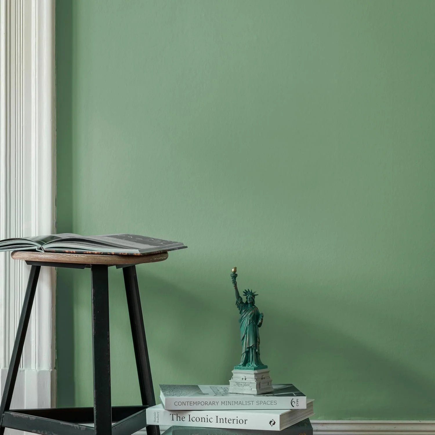 A small black side table with an open book on top, a miniature Statue of Liberty on stacked books, and a green wall in the background.