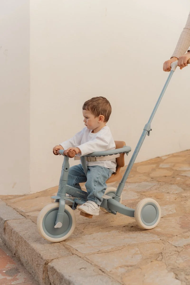 A young boy sitting in a stroller being pushed by an adult person, outdoors on a stone surface, with a plain white wall in the background.