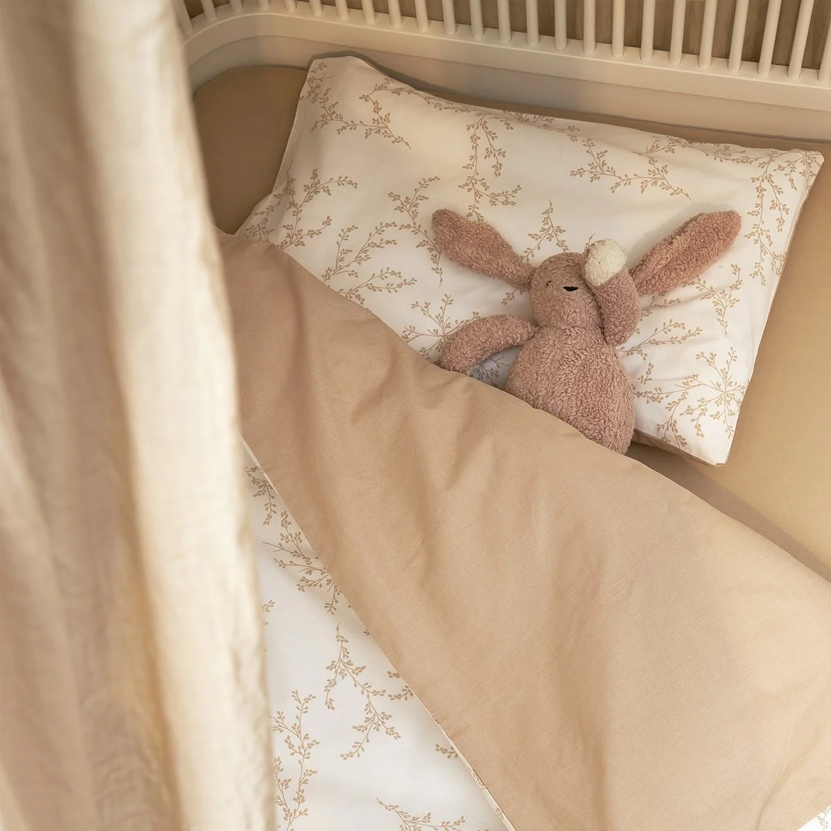 A pink plush bunny toy with a white pom-pom tail lying on a bed with beige and white sheets, white pillow with a delicate tan leafy pattern, and a beige blanket, with a white wooden headboard and cream-colored curtain partially visible.
