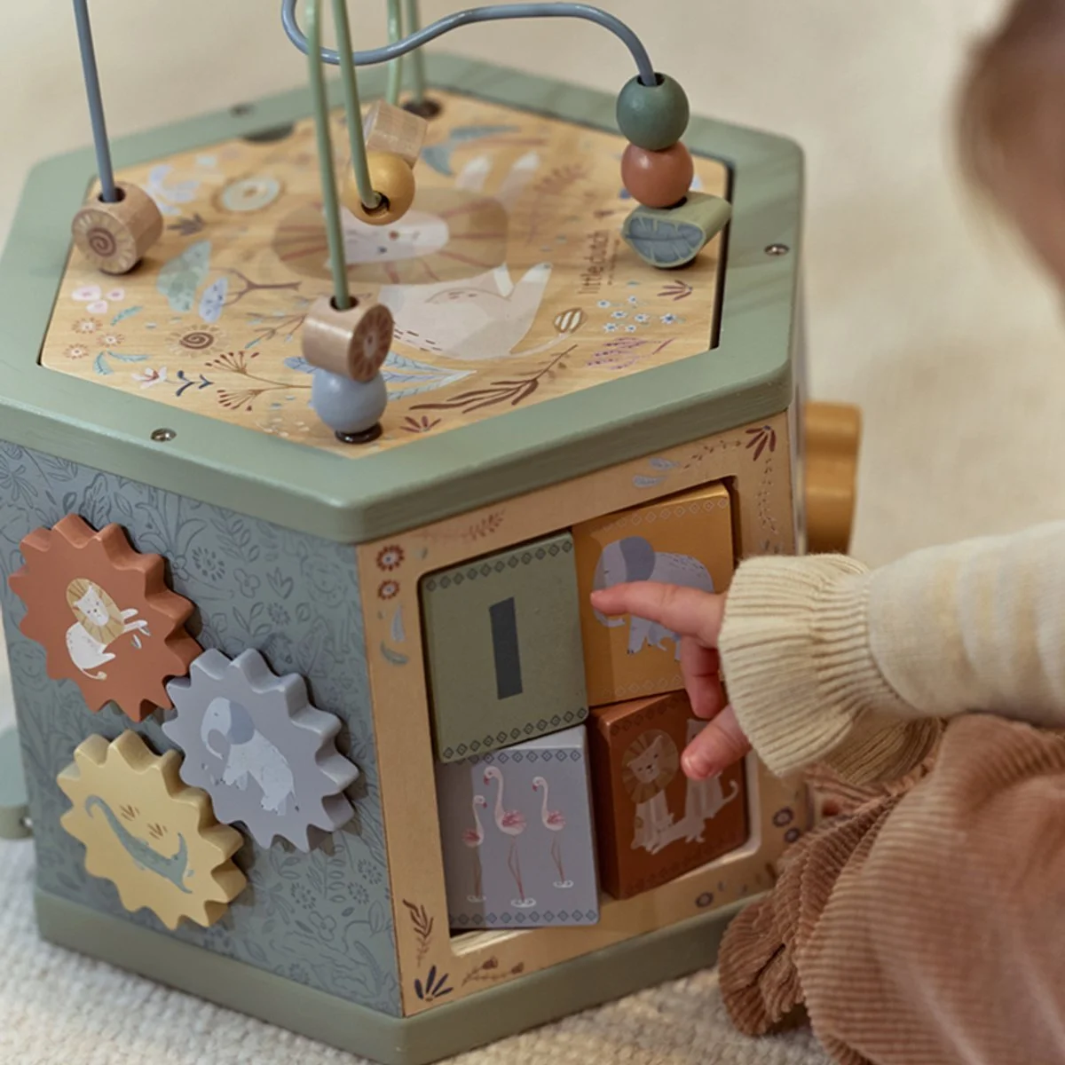 A child's hand pressing a button on a wooden activity cube with animal illustrations and gears, placed on a beige carpet.