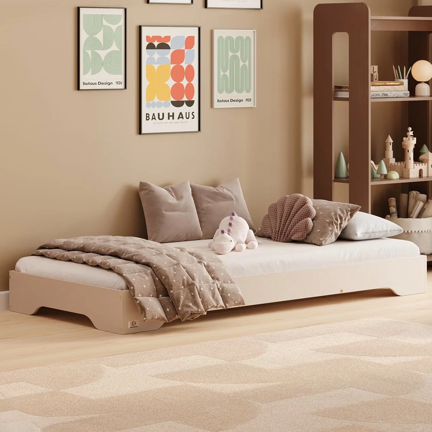 A beige bed with soft pillows and a plush toy, covered with a star-patterned blanket, in a cozy room with framed artwork on the wall, a light-colored rug, and a wooden bookshelf filled with toys and decorative items.