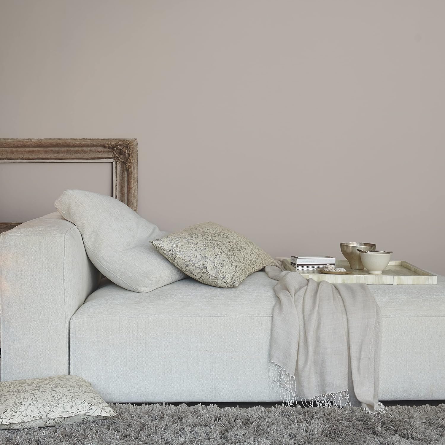 A minimalist living room scene with a white sofa, beige and patterned pillows, a tray with bowls and a book, and a shaggy gray rug.