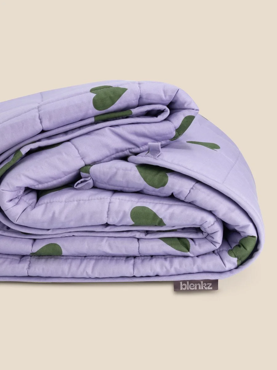 Folded lavender quilt with green leaf patterns, labeled 'blenaz,' on a beige background.
