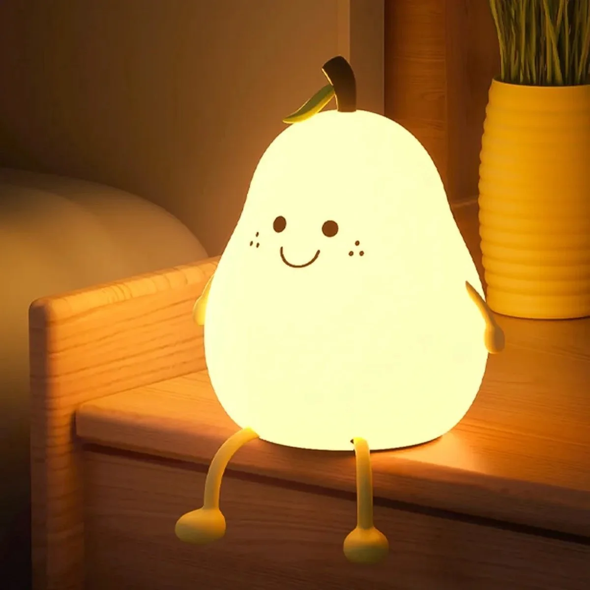 A cute pear-shaped lamp with a smiling face, tiny arms and legs, sitting on a wooden surface in a cozy room with a potted plant in the background.
