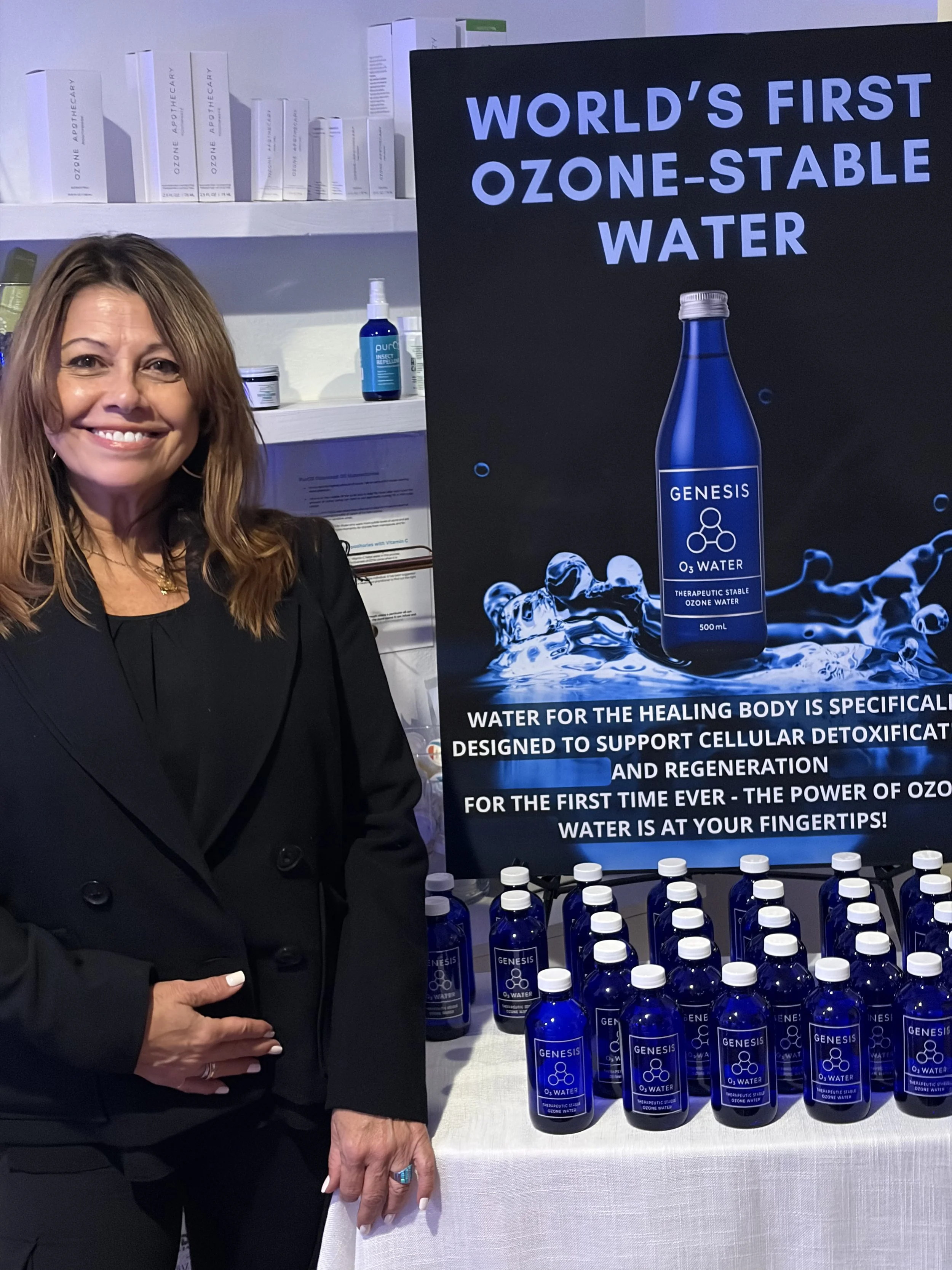 A woman standing next to a display promoting Genesis Ozone-Stable Water, with multiple bottles of the water on the table and a poster highlighting its benefits and features.