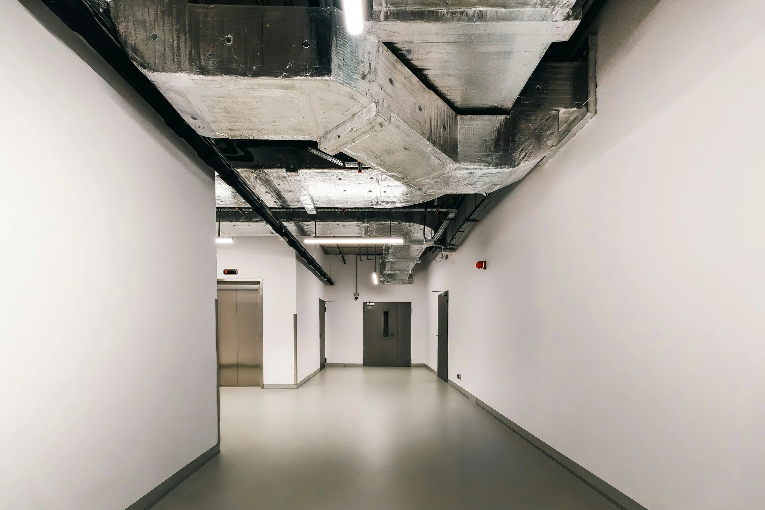Empty hallway in a modern building with white walls, gray doors, and an exposed metallic ceiling with ductwork and black pipes.