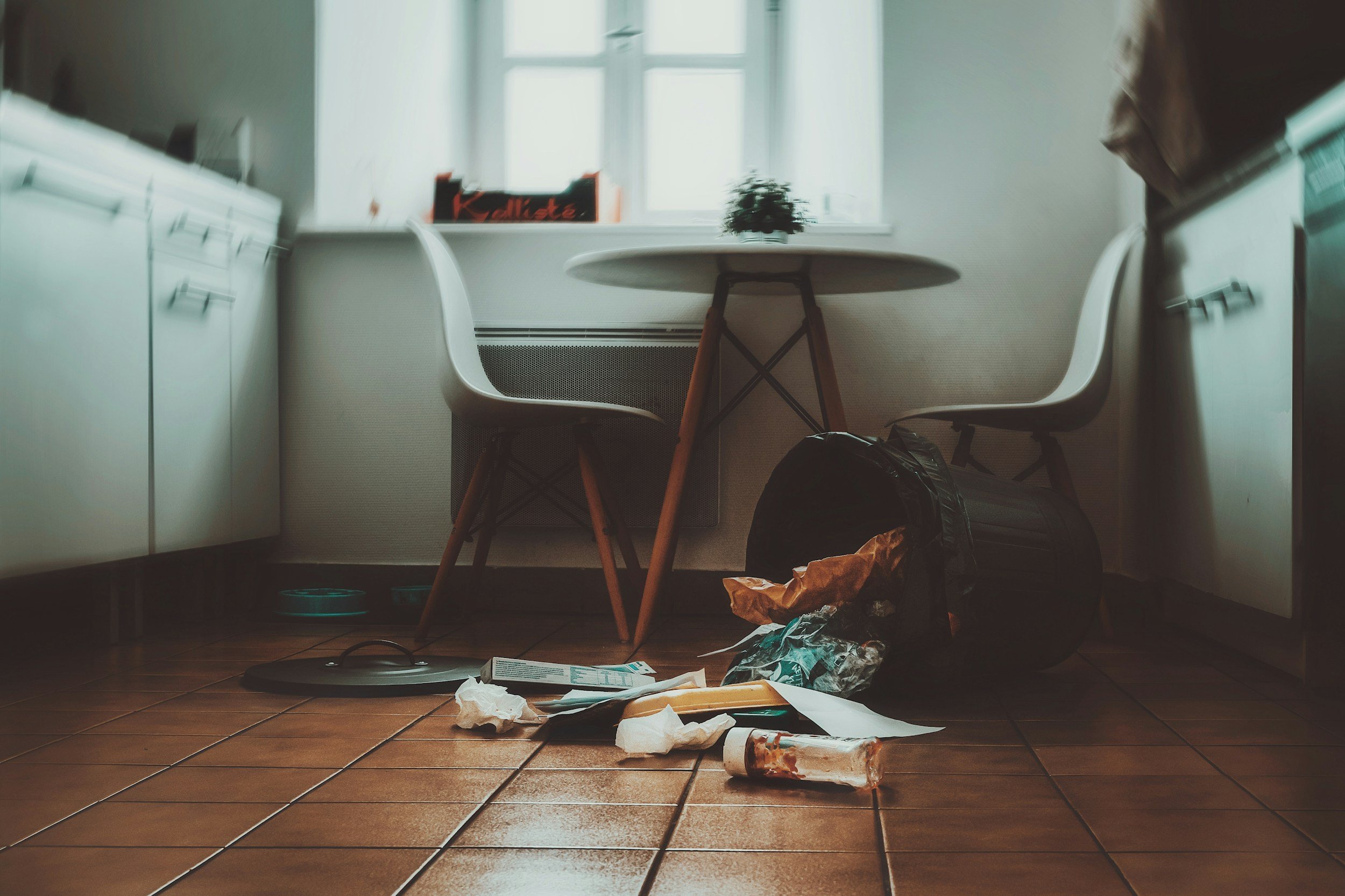 Cluttered kitchen floor with trash, food wrappers, and paper scattered around. There is a black backpack and an orange bag on the floor. A small round table with a plant on top is positioned between two white chairs, with a window in the background.