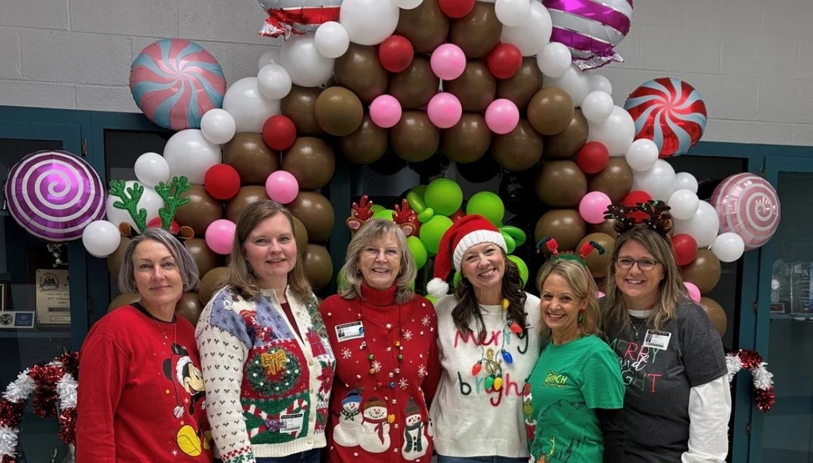 Six women wearing Christmas-themed sweaters and holiday accessories standing in front of a festive balloon arch decorated with colorful balloons and large peppermint swirl balloons.