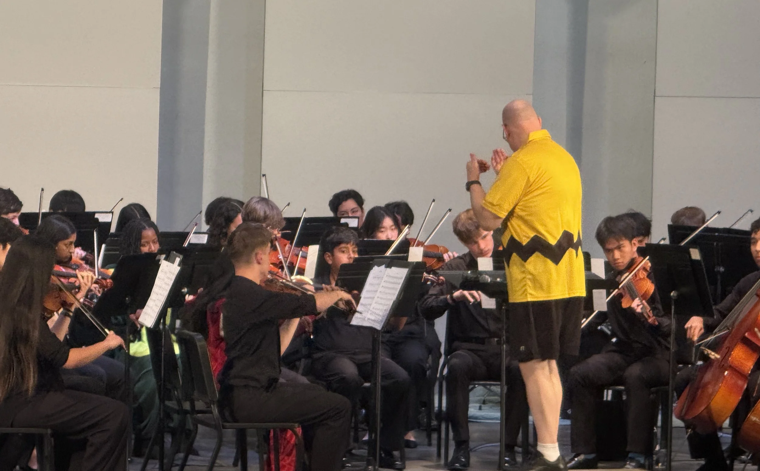 An orchestra conductor in a yellow shirt leading a group of young musicians playing string instruments on stage.