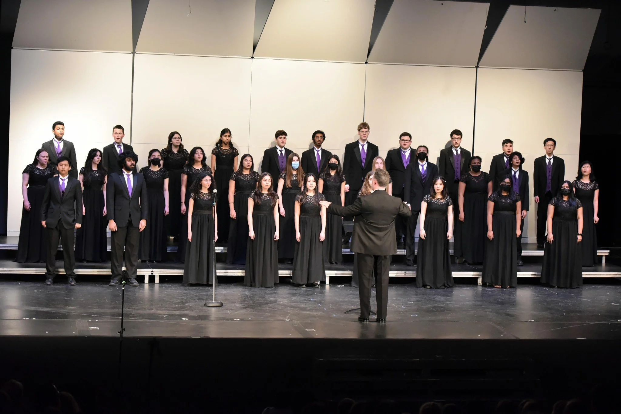 A choir of young men and women singing on a stage, conducted by a conductor in a black suit, with some choir members wearing black masks.