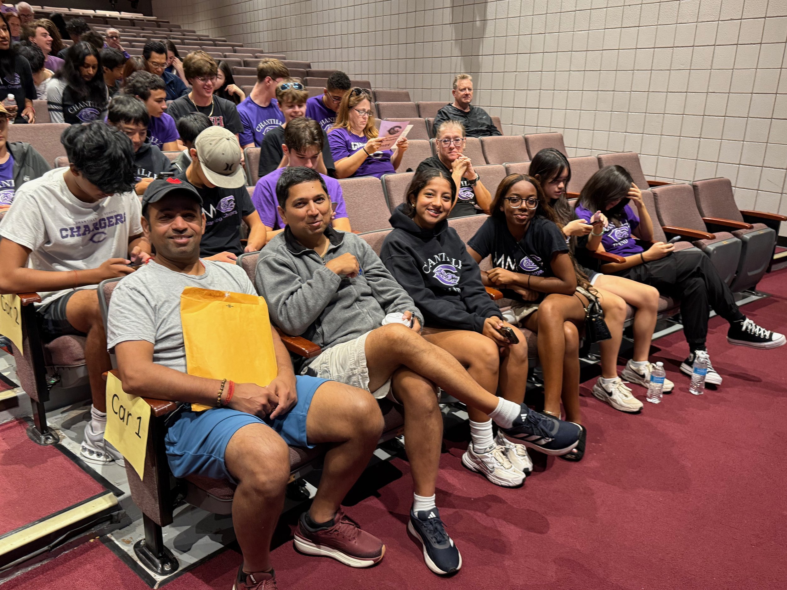 Group of people sitting in an auditorium, with some smiling and others engaged in reading or using their phones. The front row has five individuals, with young students wearing black and purple shirts, and an adult man holding a yellow paper bag. The seats are labeled, and there are water bottles on the floor near some of the people.