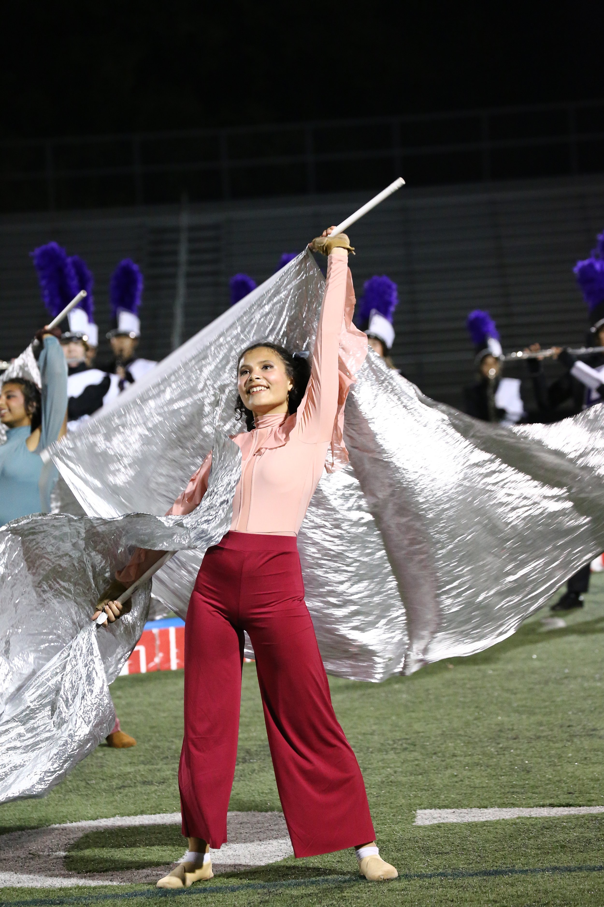 A young woman in a pink top and red pants performing with a silver flag on a football field during a nighttime marching band performance, with band members in uniform and purple plumed hats in the background.