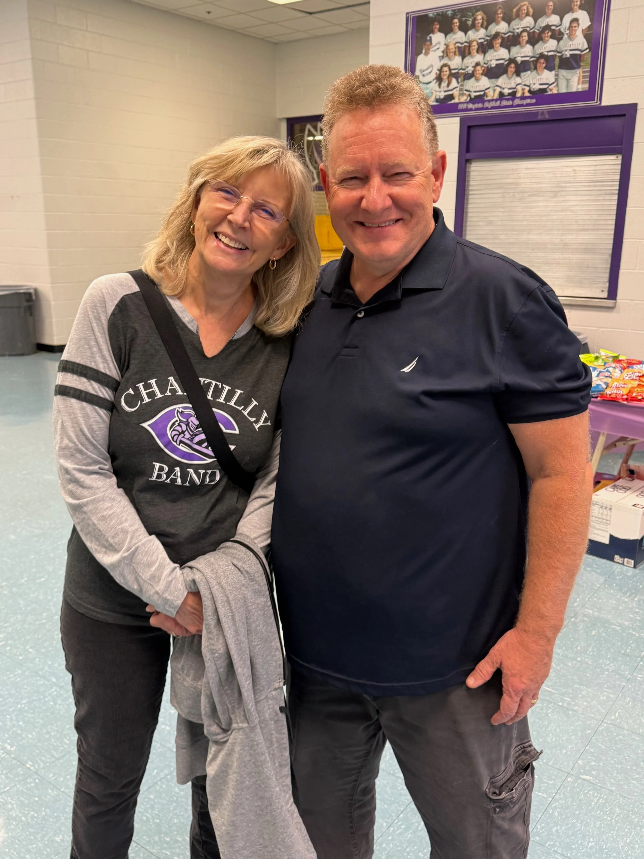 A smiling woman and man posing together indoors, woman wearing a T-shirt with 'CHARLOTTE BAND' logo, woman has blonde hair and glasses, man has short curly hair, both are smiling.