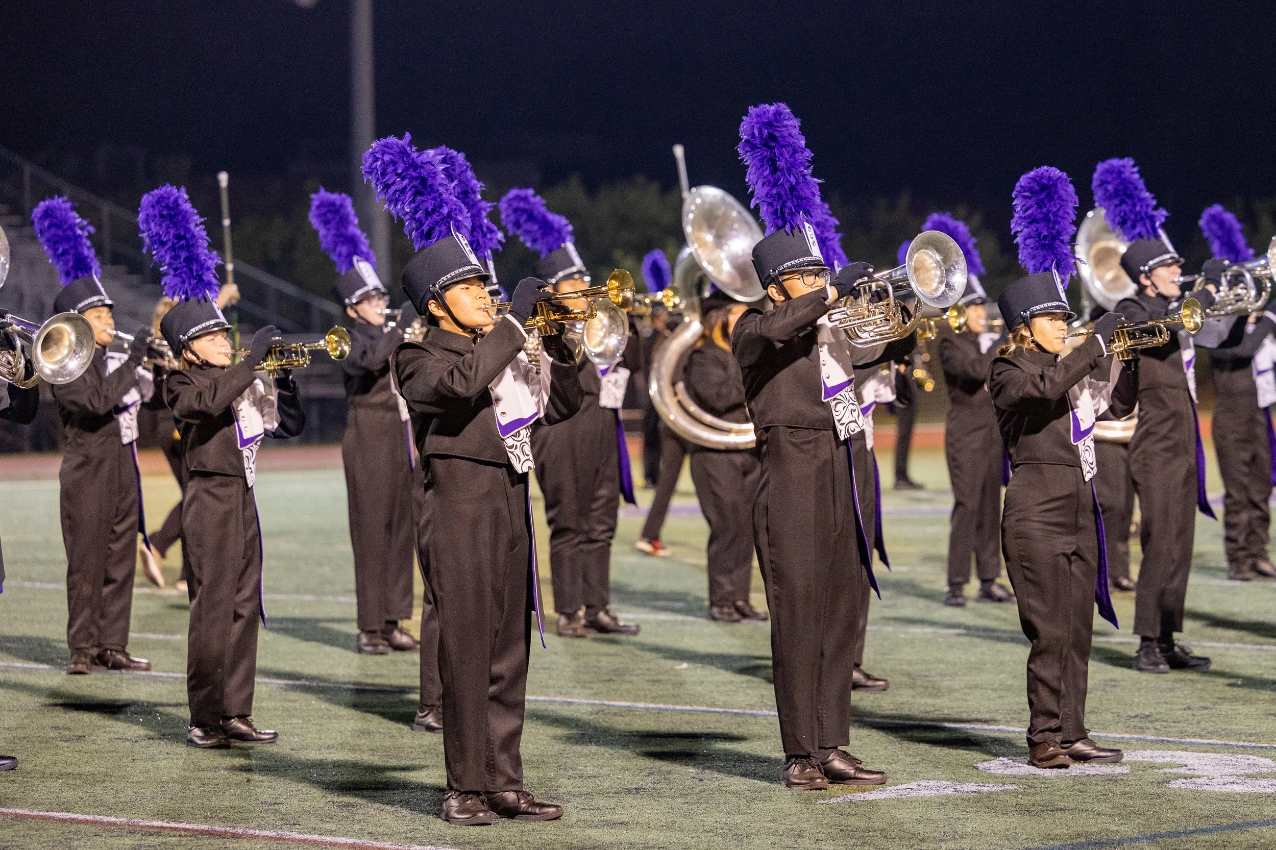 High school marching band performing on a football field at night, dressed in black uniforms with purple feathers on their hats.