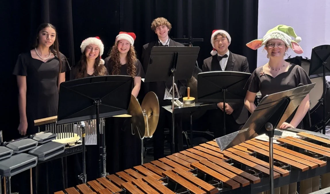 Group of six young musicians in holiday costumes, some wearing Santa hats and one with a novelty elf hat, standing behind musical instruments in a performance setting.