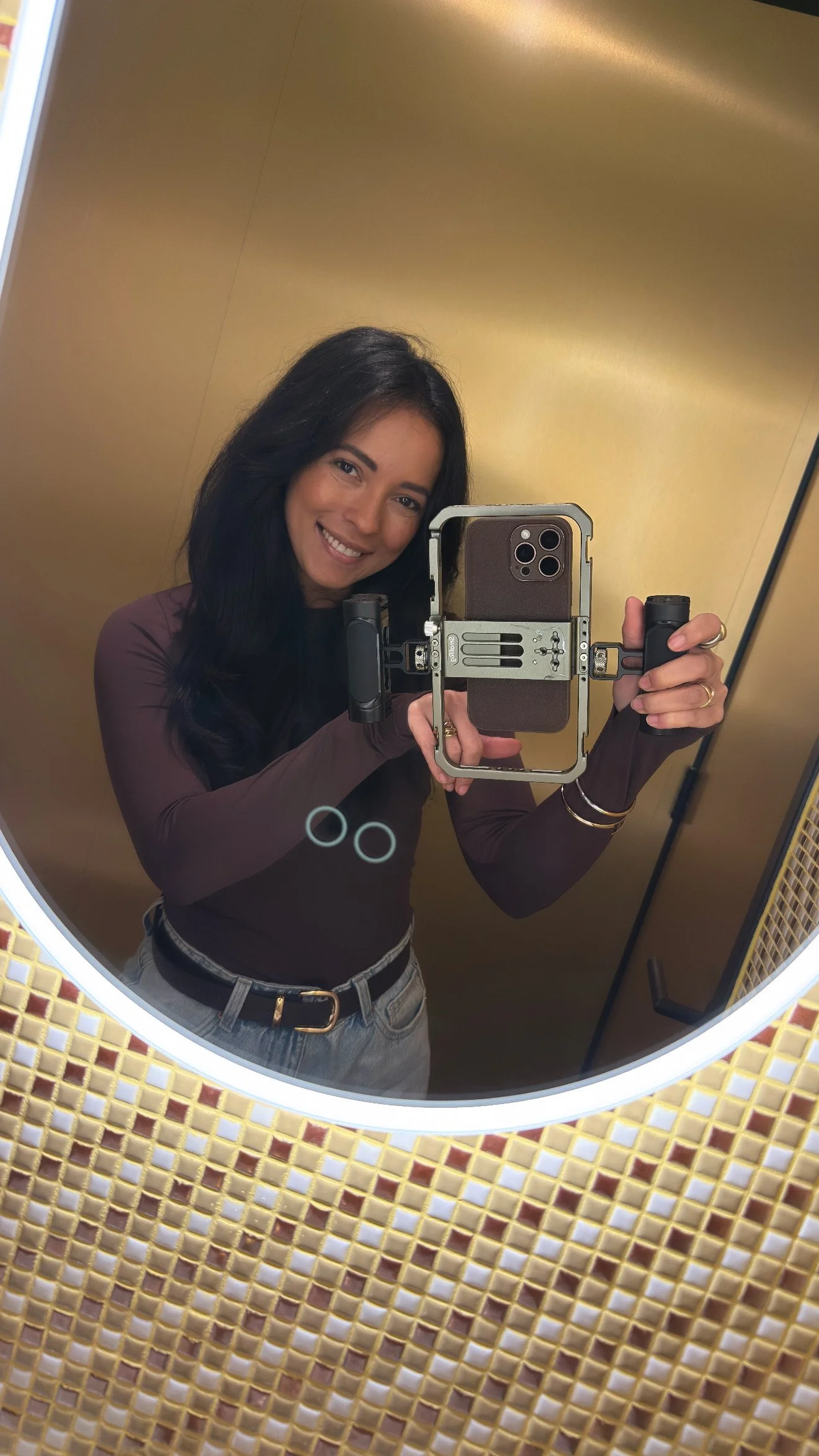 Vrouw met lange donkere haar in spiegel of camera, glimlachend, in een kamer met gouden muur, gekleed in een bruin shirt en lichtgrijze broek, holding een camera of stabilizer.