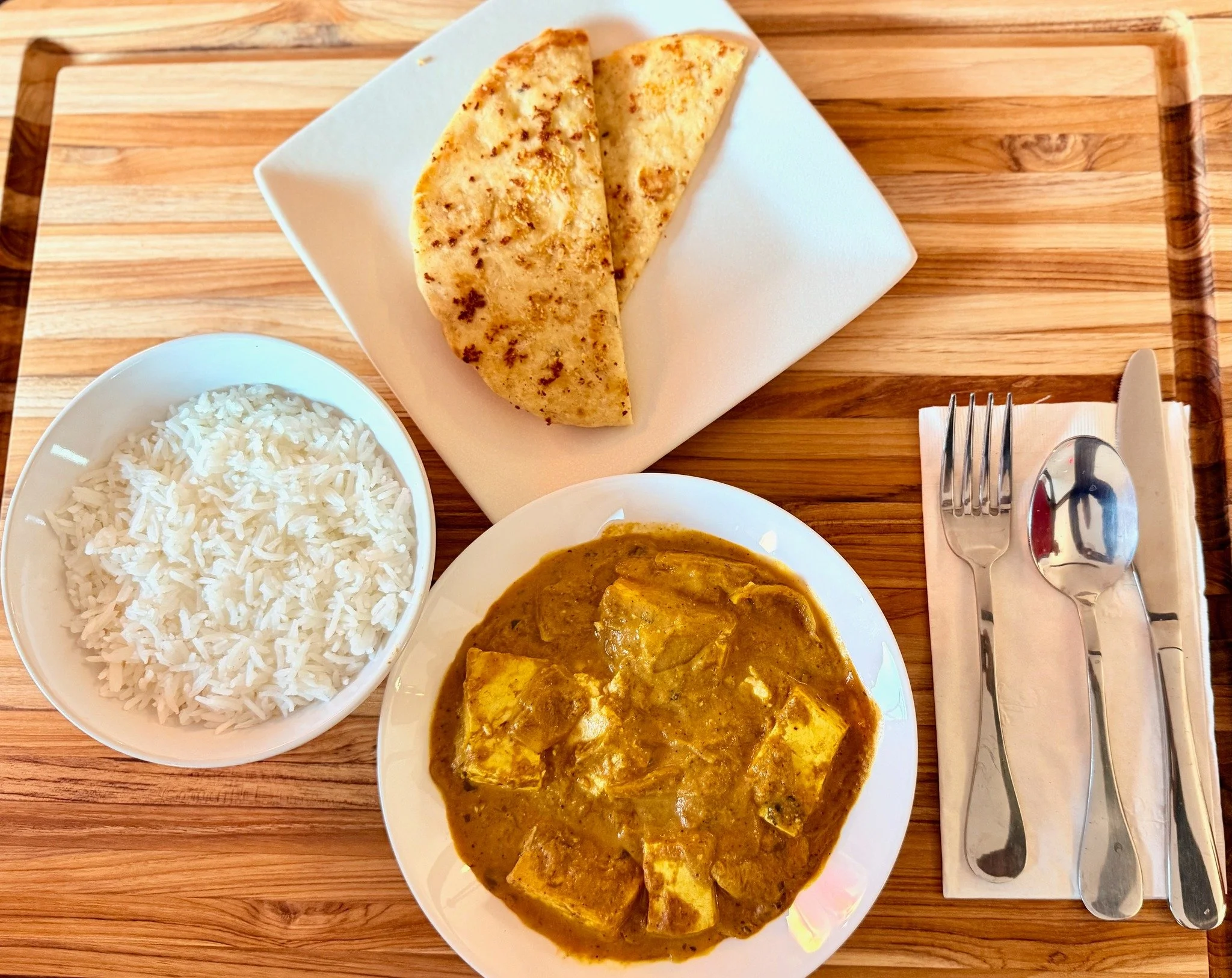 Indian meal on a wooden tray includes a bowl of white rice, a plate of tofu curry in brown sauce, and two pieces of fried flatbread on a white square plate.