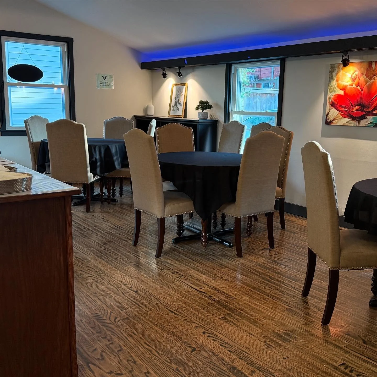A dining room with several round tables covered with black tablecloths and beige upholstered chairs. There are two windows, artwork on the walls, and decorative plants.