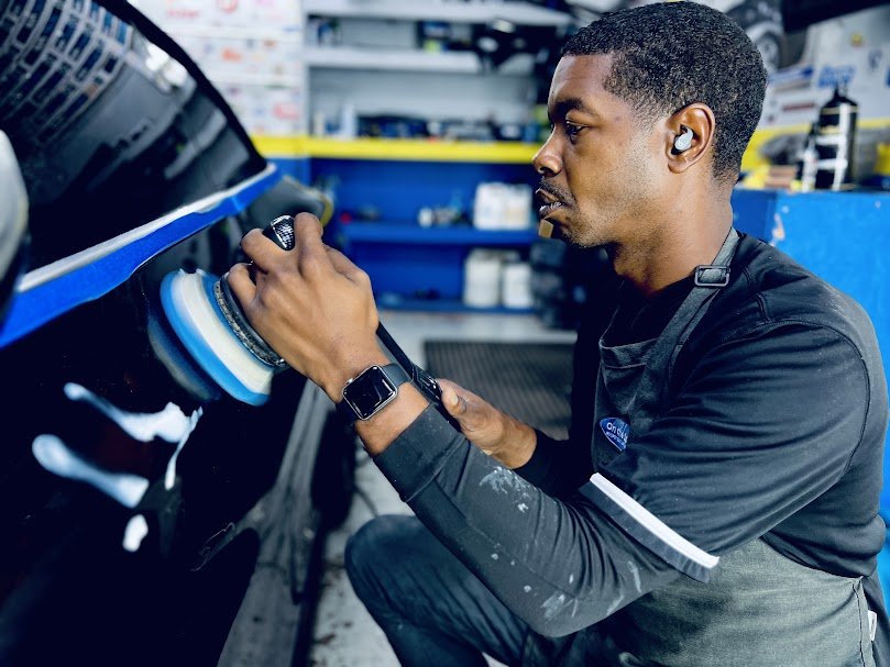 A man wearing a black work shirt with a watch on his left wrist is polishing a car's black exterior with a blue and white buffer tool in a workshop.