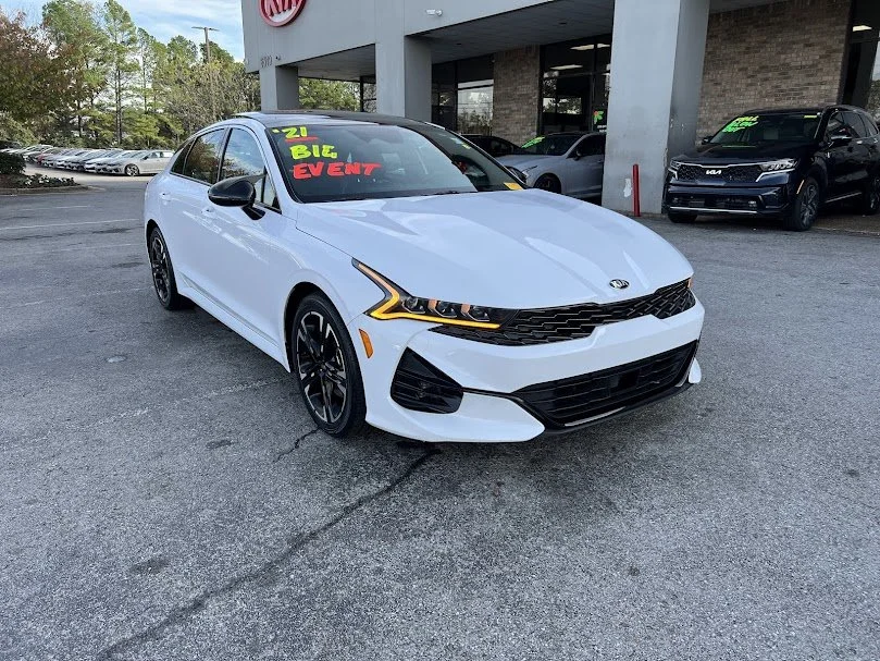 White Kia sedan parked in a dealership lot, with a sign in the windshield indicating a 2021 model and an upcoming event.