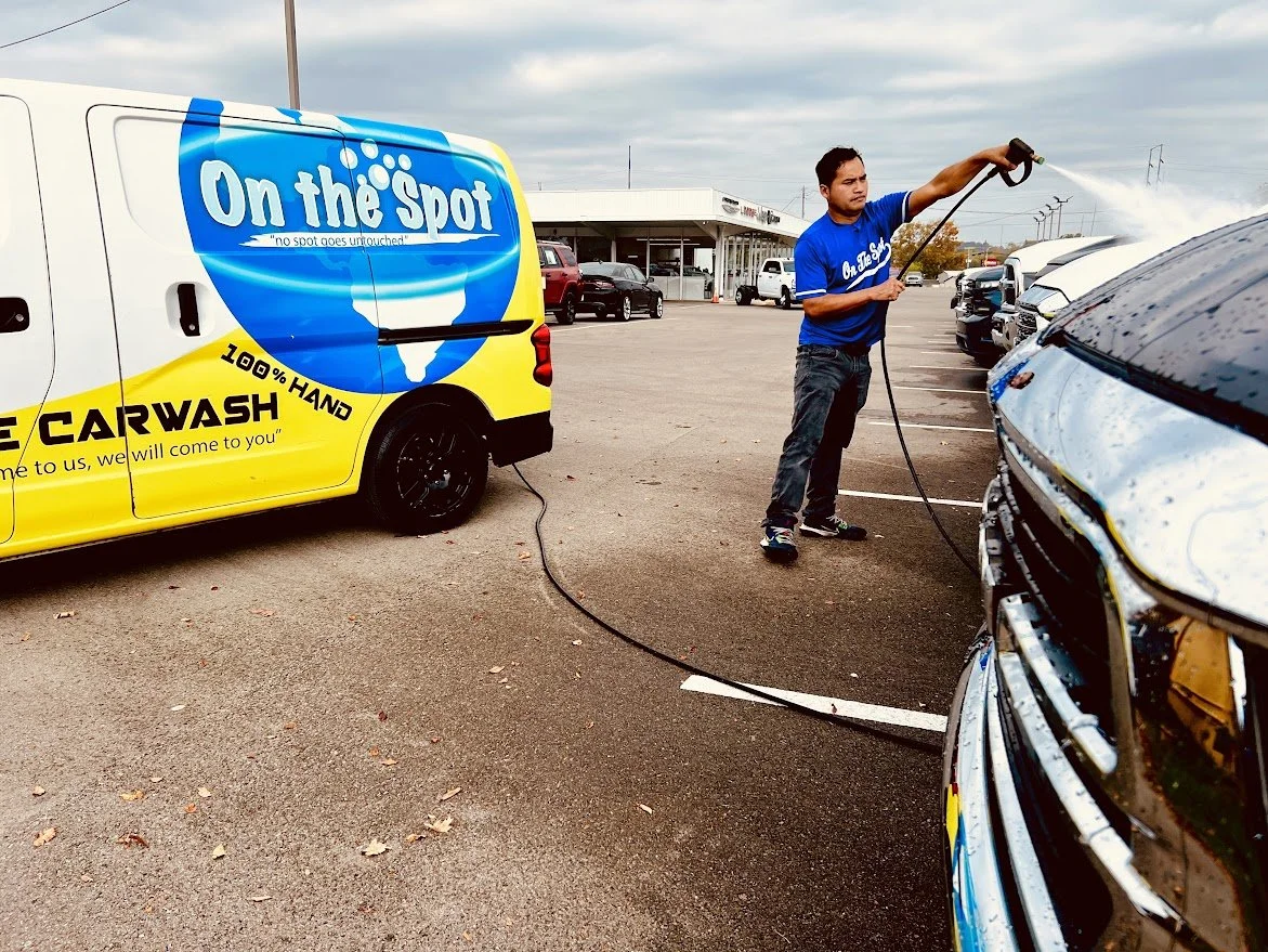 A man in a blue shirt is using a pressure washer to clean the front of a vehicle at a car wash. The car wash van with bright yellow and blue colors is nearby, with the text "On the Spot" and advertising "100% Hand" car wash. The scene is in a parking lot with several parked cars and a building in the background.