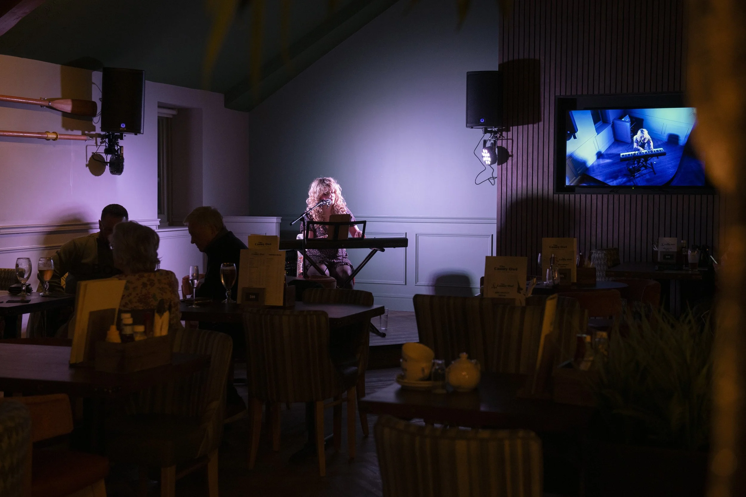 A live musician performing on a stage. She's playing the keyboard and singing in front of tables with people who are having drinks.