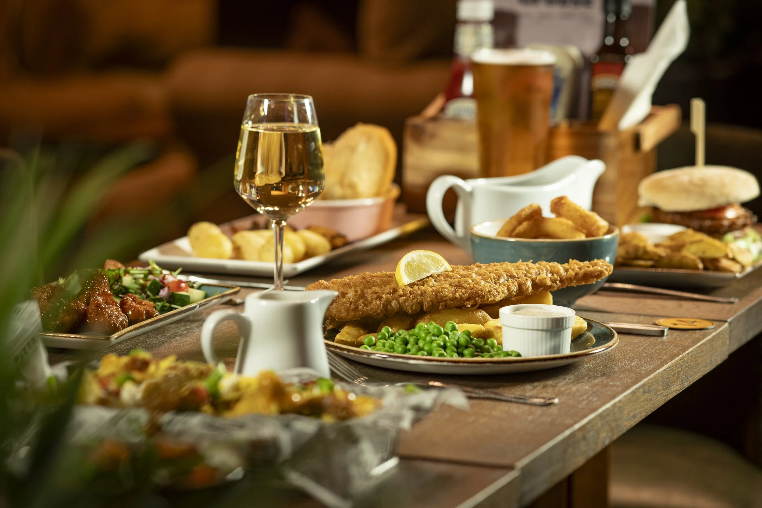 Table set up with multiple food dishes including fish and chips with peas, BBQ cauliflower bites, veggie burger with chips. There's a glass of white wine and a pint of beer between the foods as well.