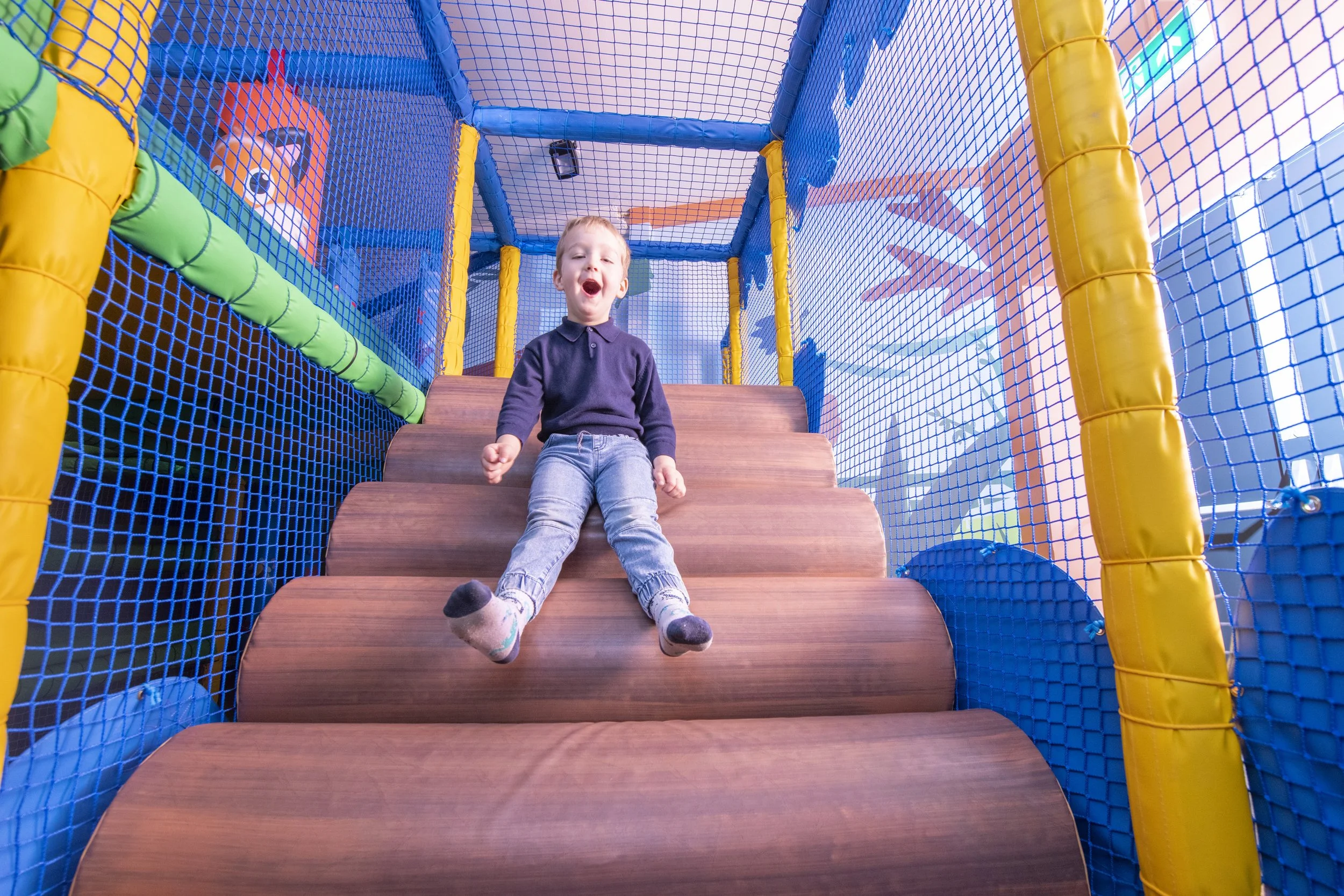 Young boy playing on indoor jungle gym with slides and padded surfaces, smiling and having fun.