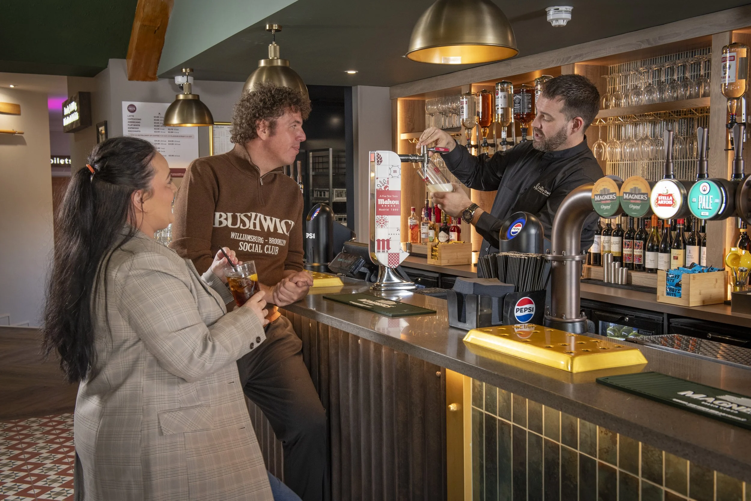 A man and woman sitting at a bar, chatting and smiling, waiting for a pint. The bar is made of wood with green chairs. Behind them, there is a bar shelf stocked with various bottles of alcohol and glassware.