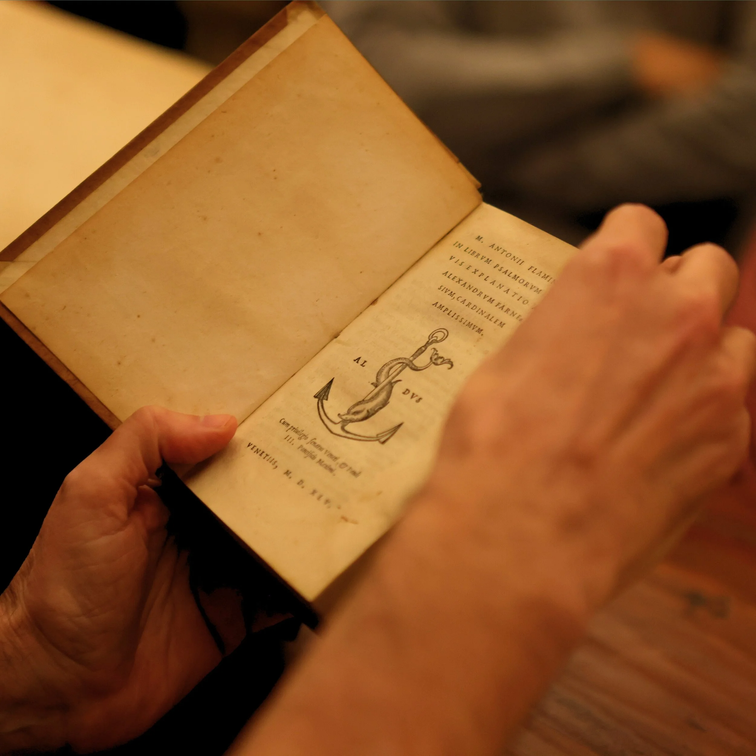 A person holding an open old book with yellowed pages, showing Latin text and a decorative illustration of an anchor.