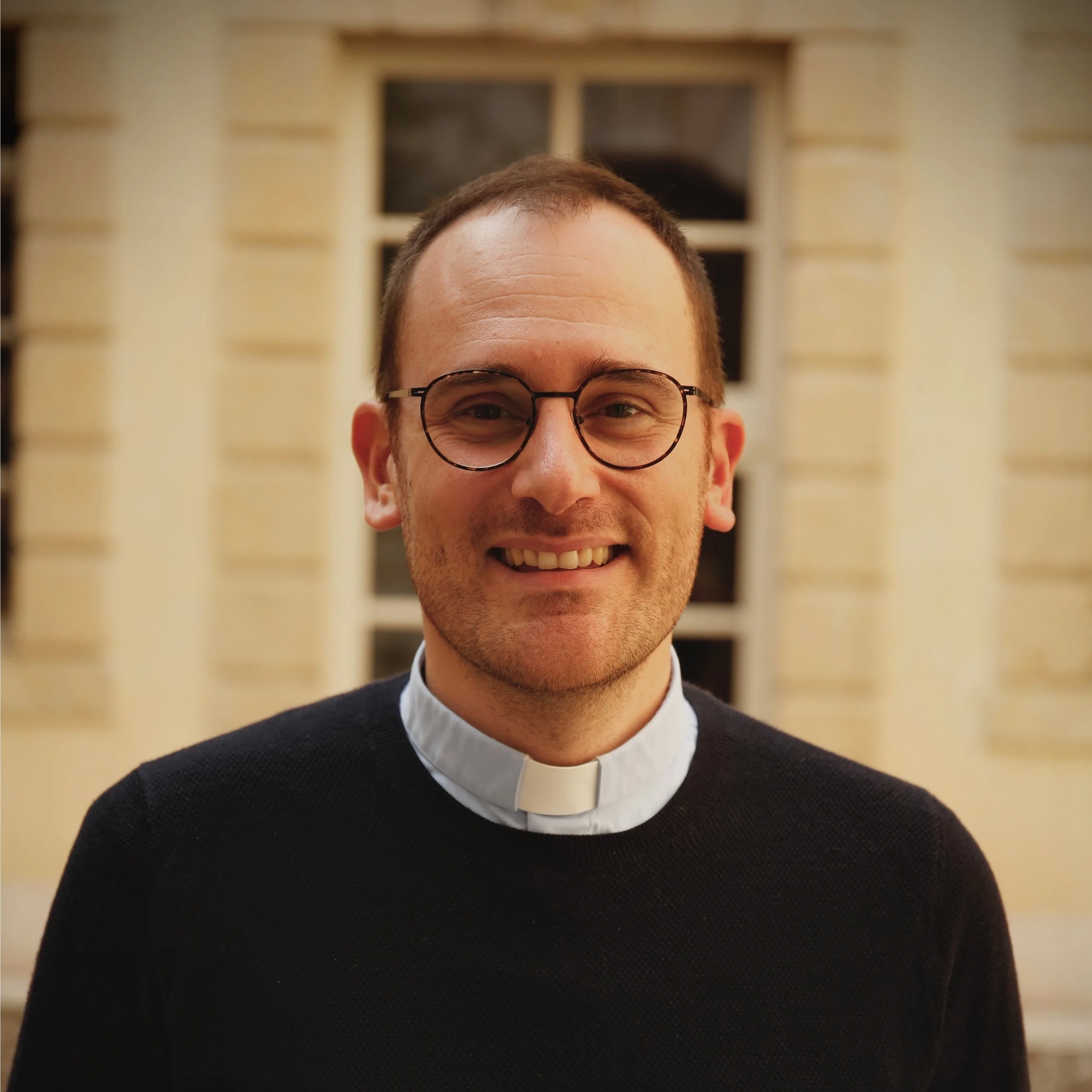 Close-up of a smiling man with glasses, wearing clerical clothing, against a blurred background of a building with windows.