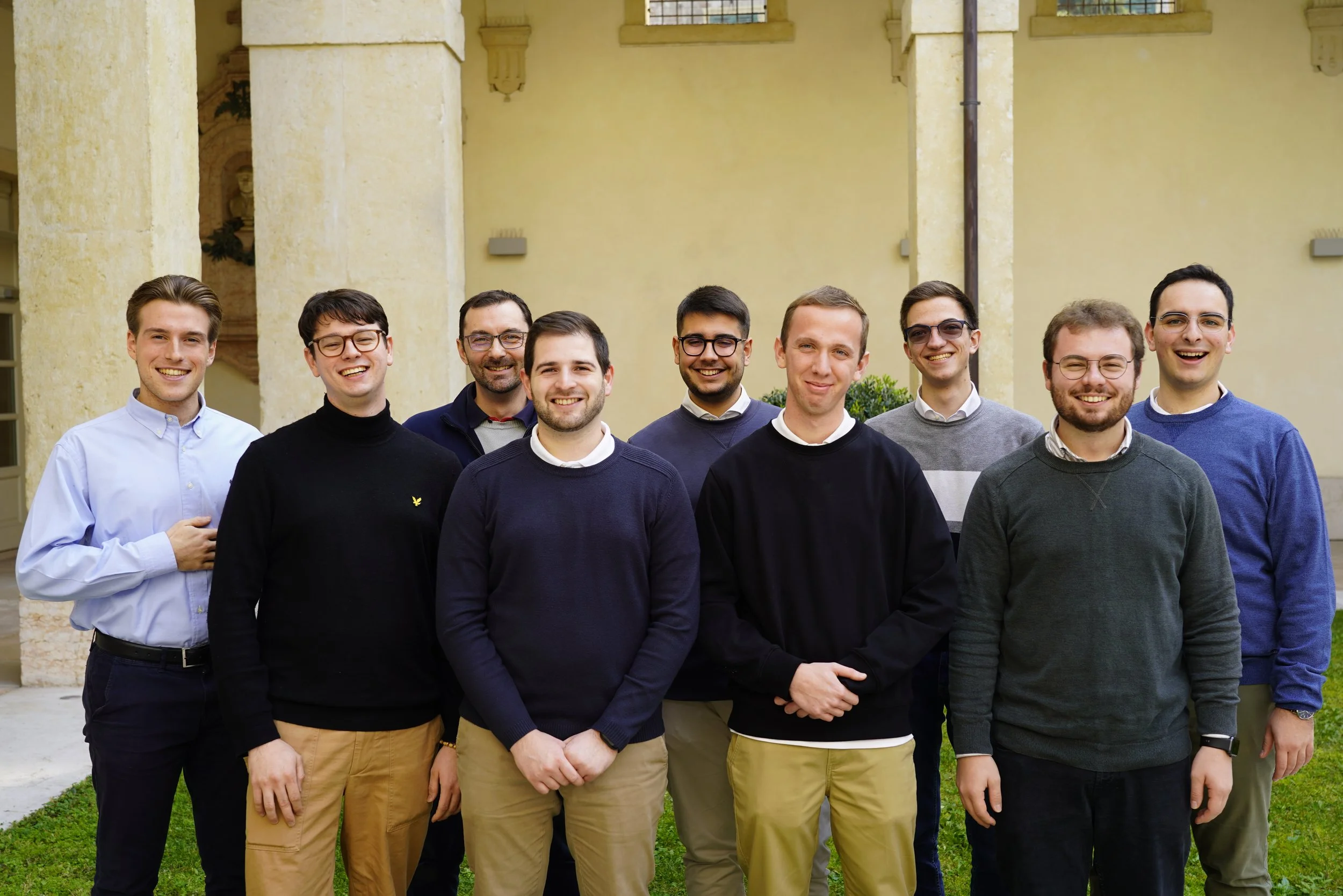 A group of ten young men standing outdoors, smiling, in front of a yellow building with columns and greenery.
