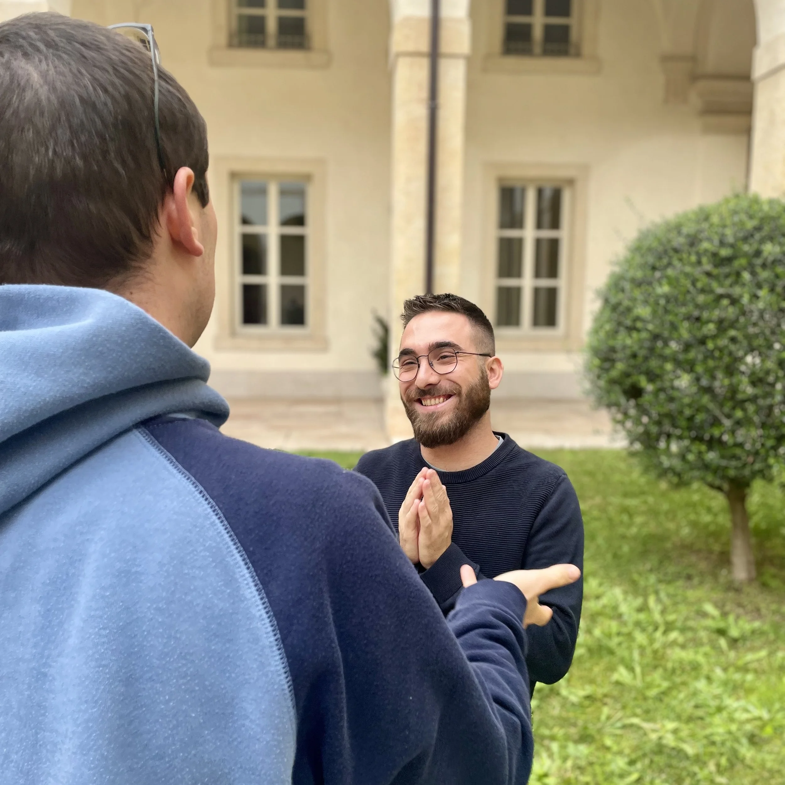 Two men are standing outdoors in front of a yellow building with large windows, engaging in a conversation. One man is wearing glasses and a dark shirt, smiling, while the other has short hair and is wearing a hoodie.