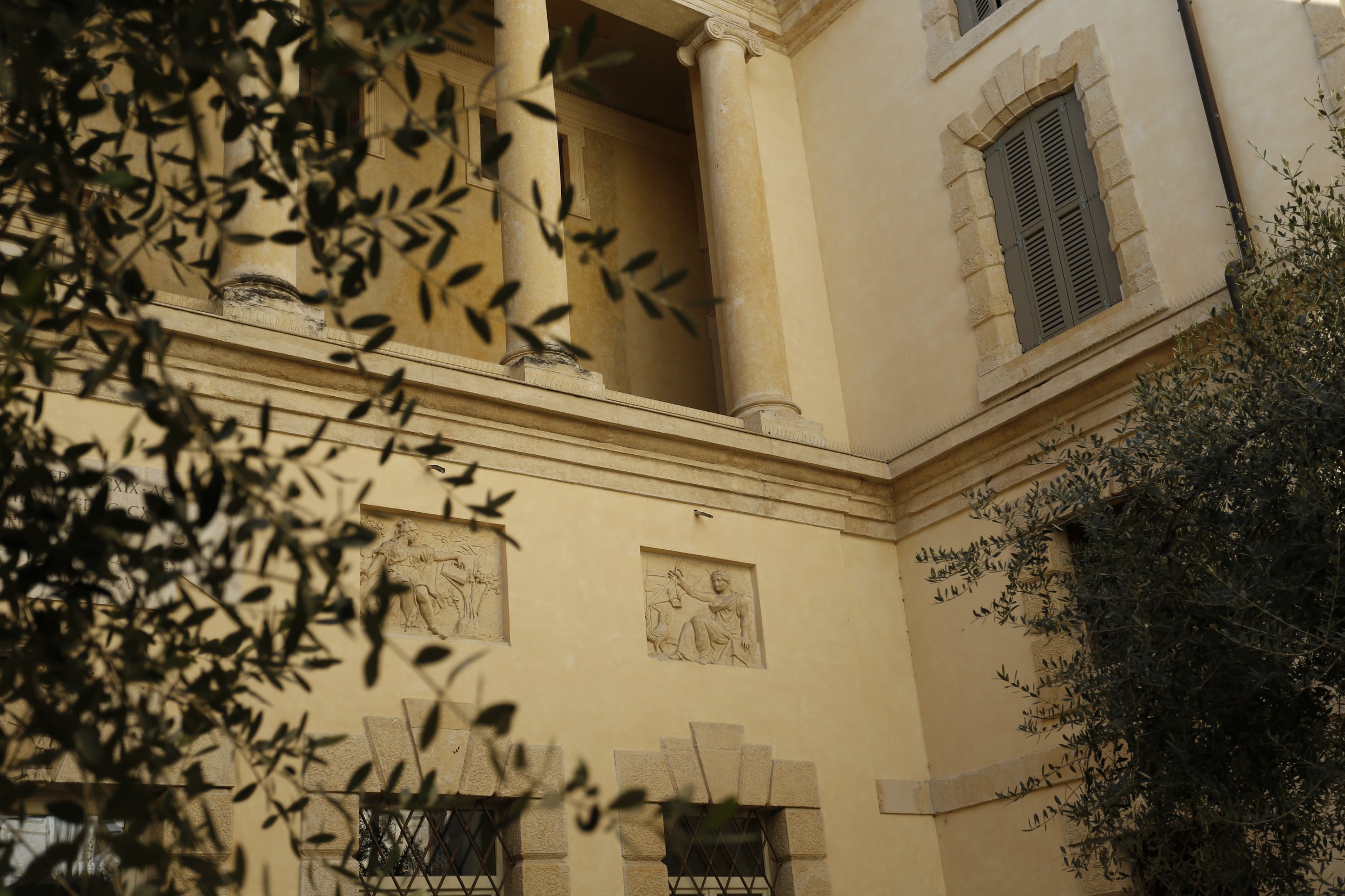 The image shows a section of a classical style building's exterior wall with beige stone or stucco finish, decorative relief sculptures depicting historical or mythological scenes, a window with closed shutters, and stone columns. There are also some dark metal window bars and surrounding greenery partially obscuring the view.