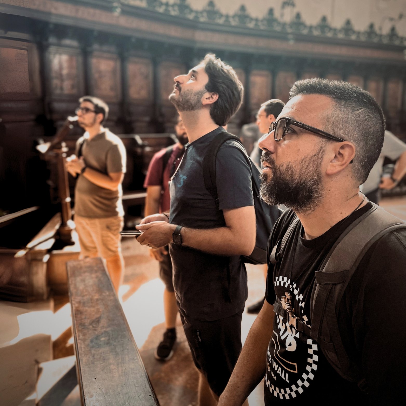 Group of men standing inside a building with wood-paneled walls, some with backpacks, looking upwards and observing something.