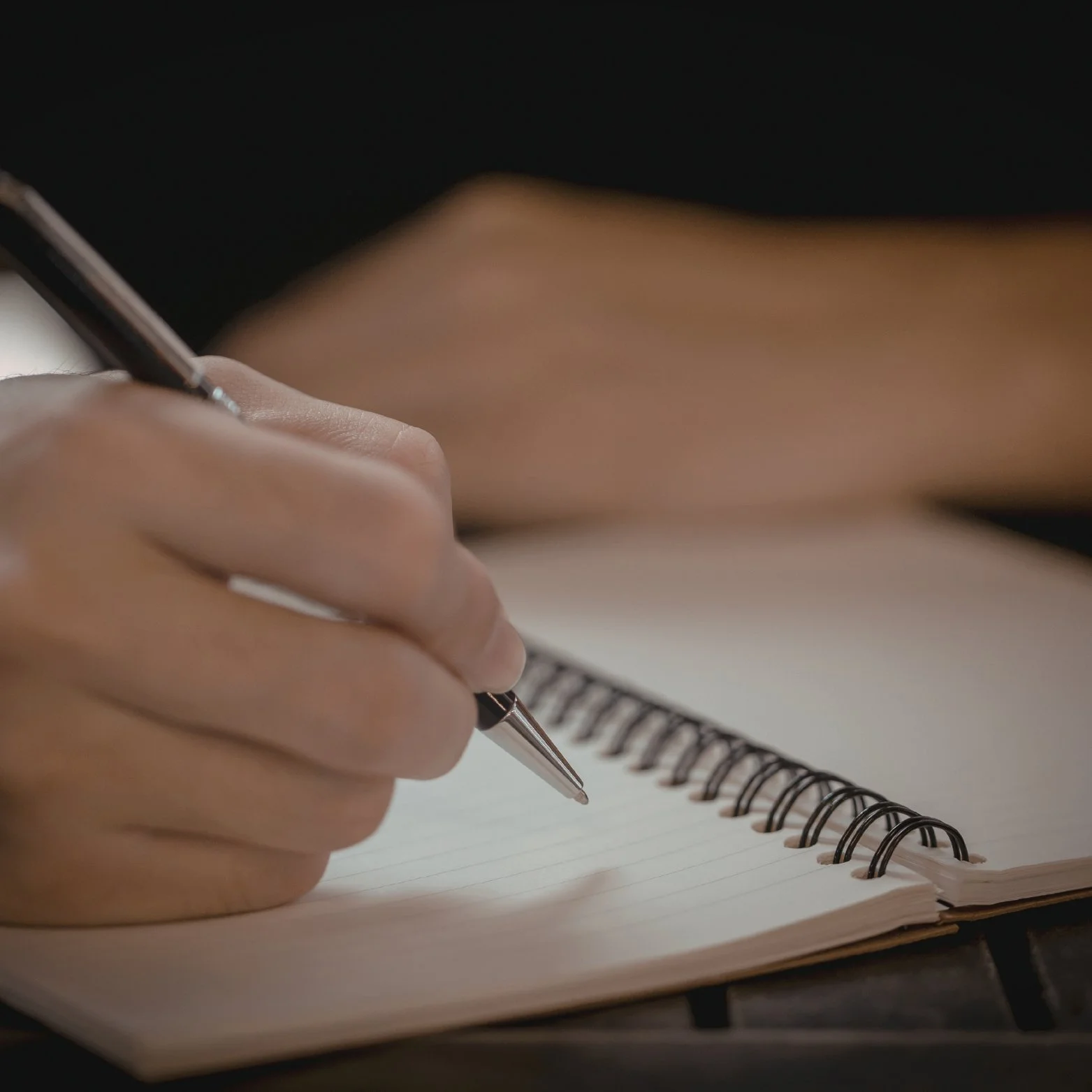 A person holding a pen and writing in a spiral notebook with lined pages.