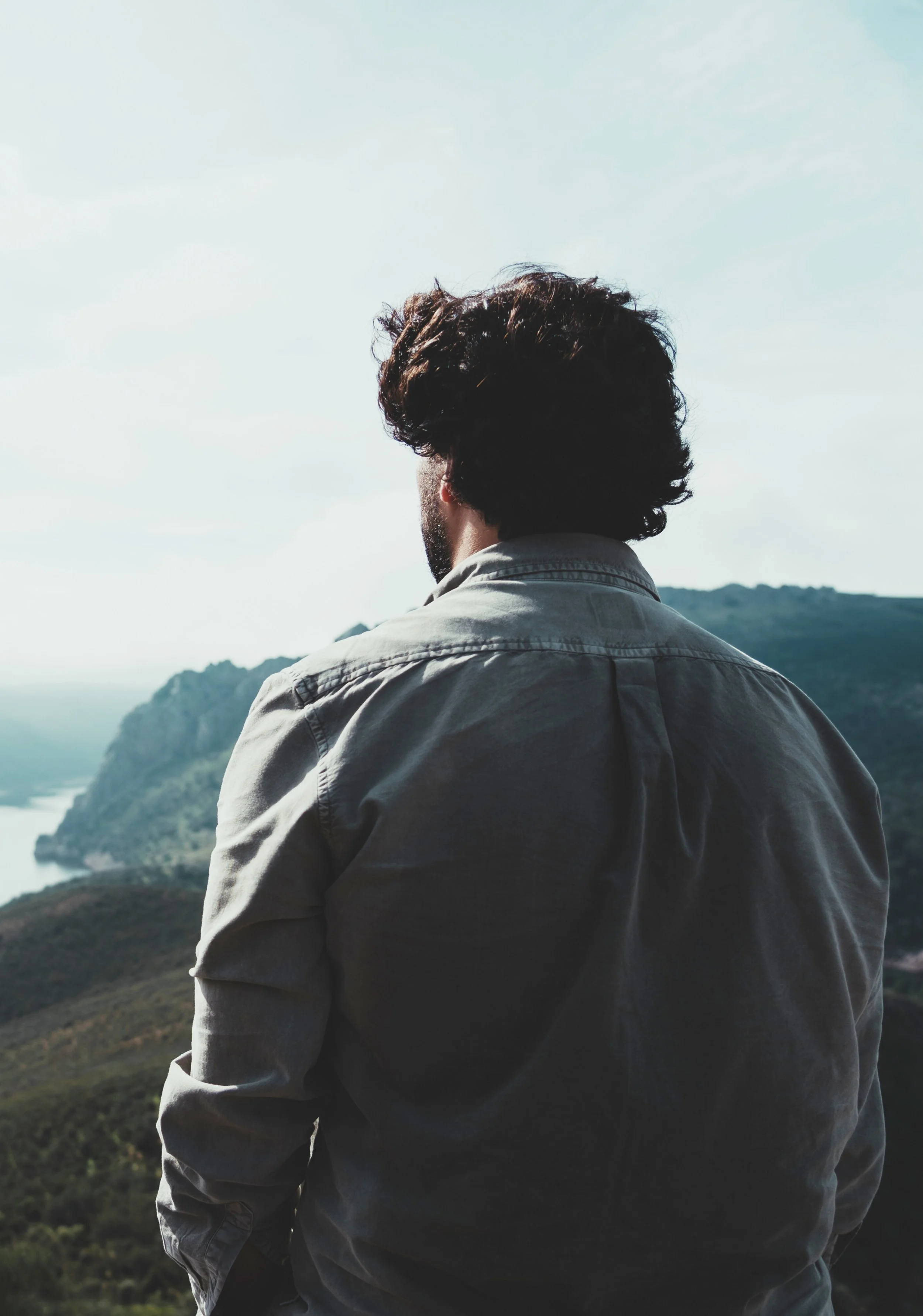 Man with curly hair and beard wearing a jacket, looking at scenic landscape of mountains and water with a cloudy sky.